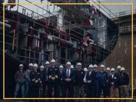Construction workers and officials wearing helmets in front of a large industrial ship under constru.